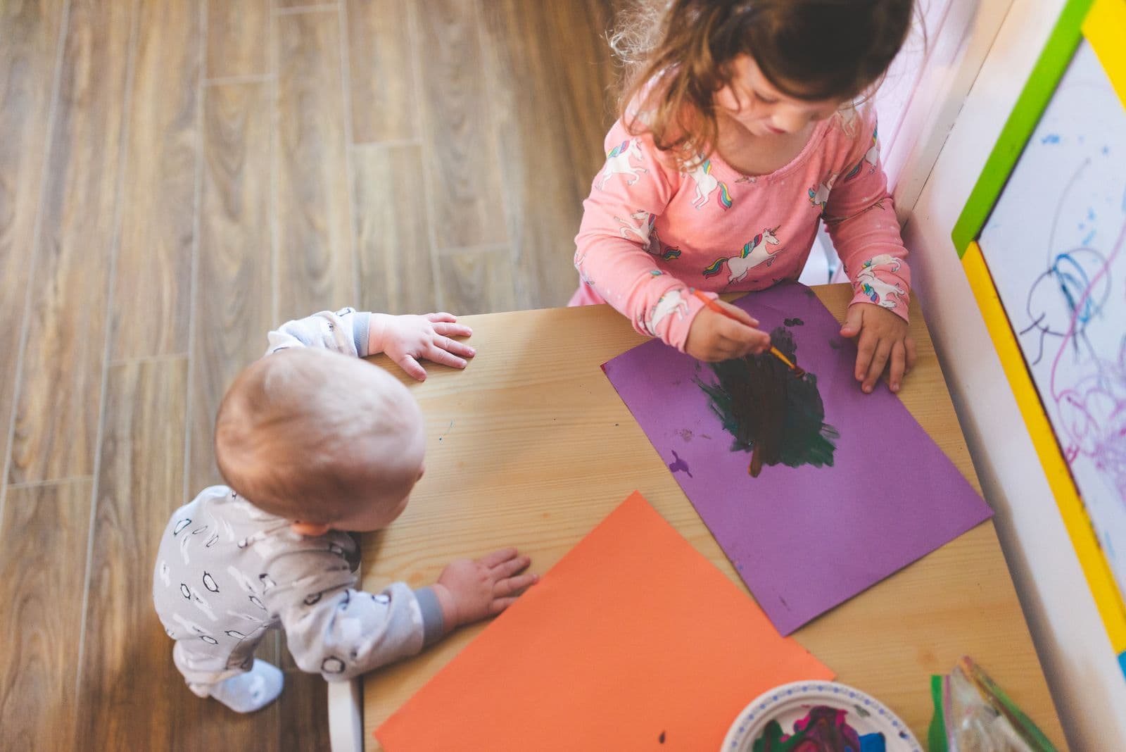 A baby kneeling at a low table, drawing on a large sheet of coloured paper.