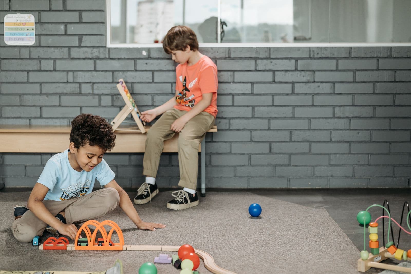 Two boys playing with wooden toys on a bench outdoors beside a brick wall.