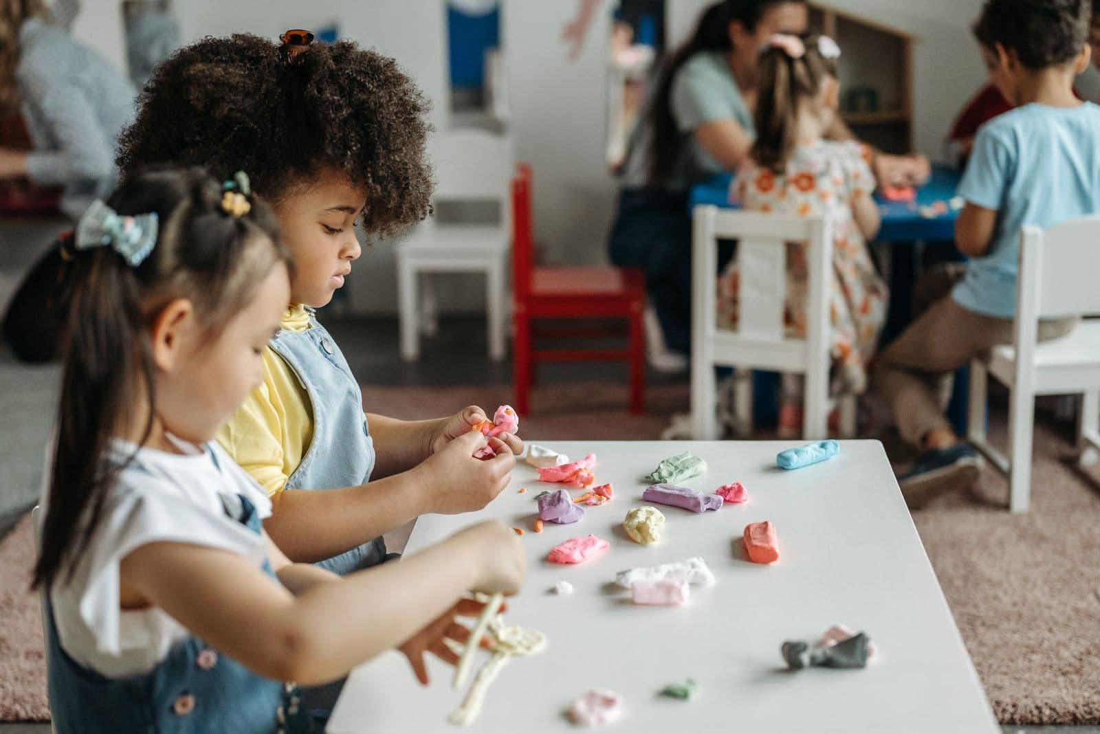 Two children shaping coloured playdough at a low table in a bright day care room, with other children and an educator working behind them.