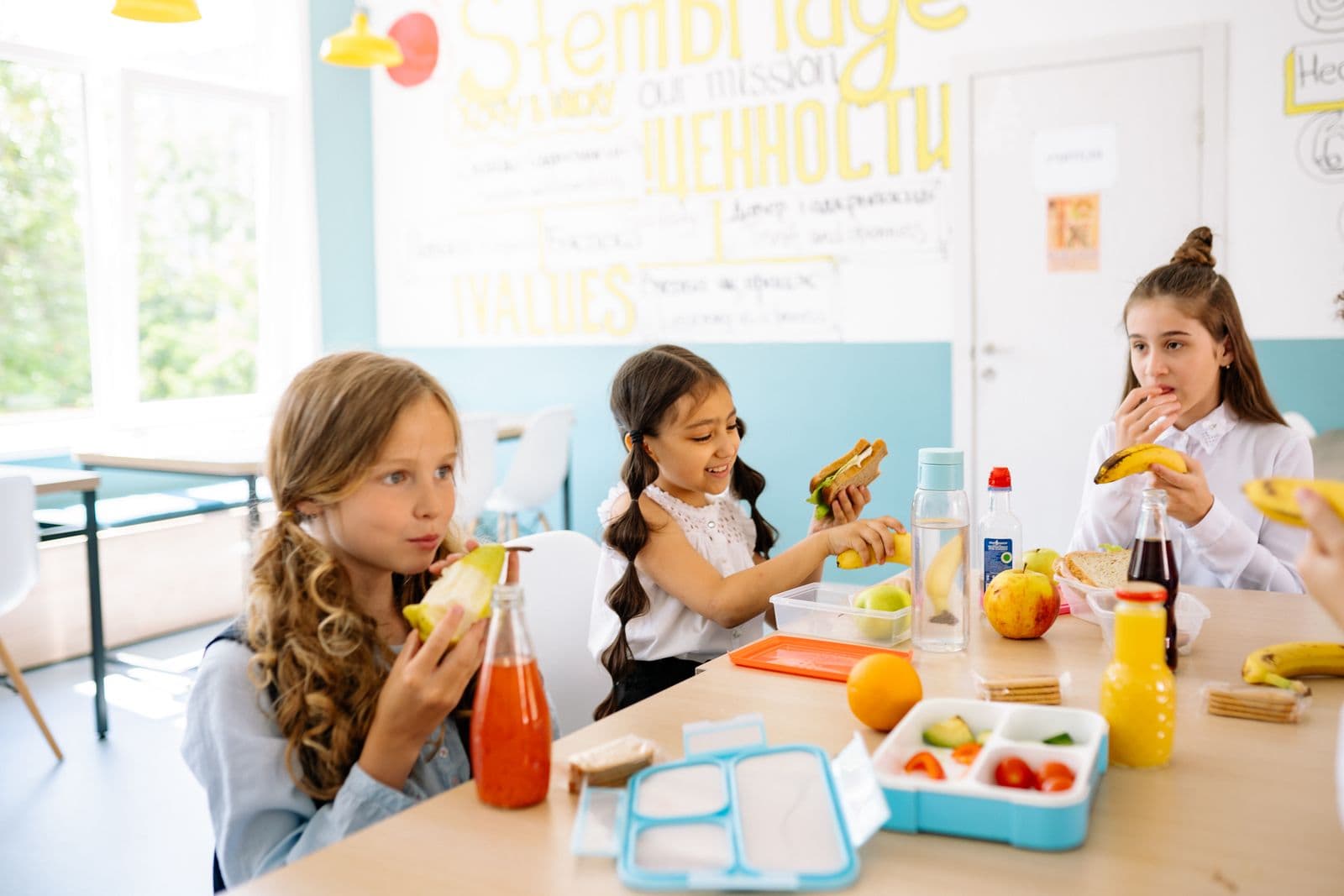 Children sitting together at a table with fruit, sandwiches and drinks in front of them.