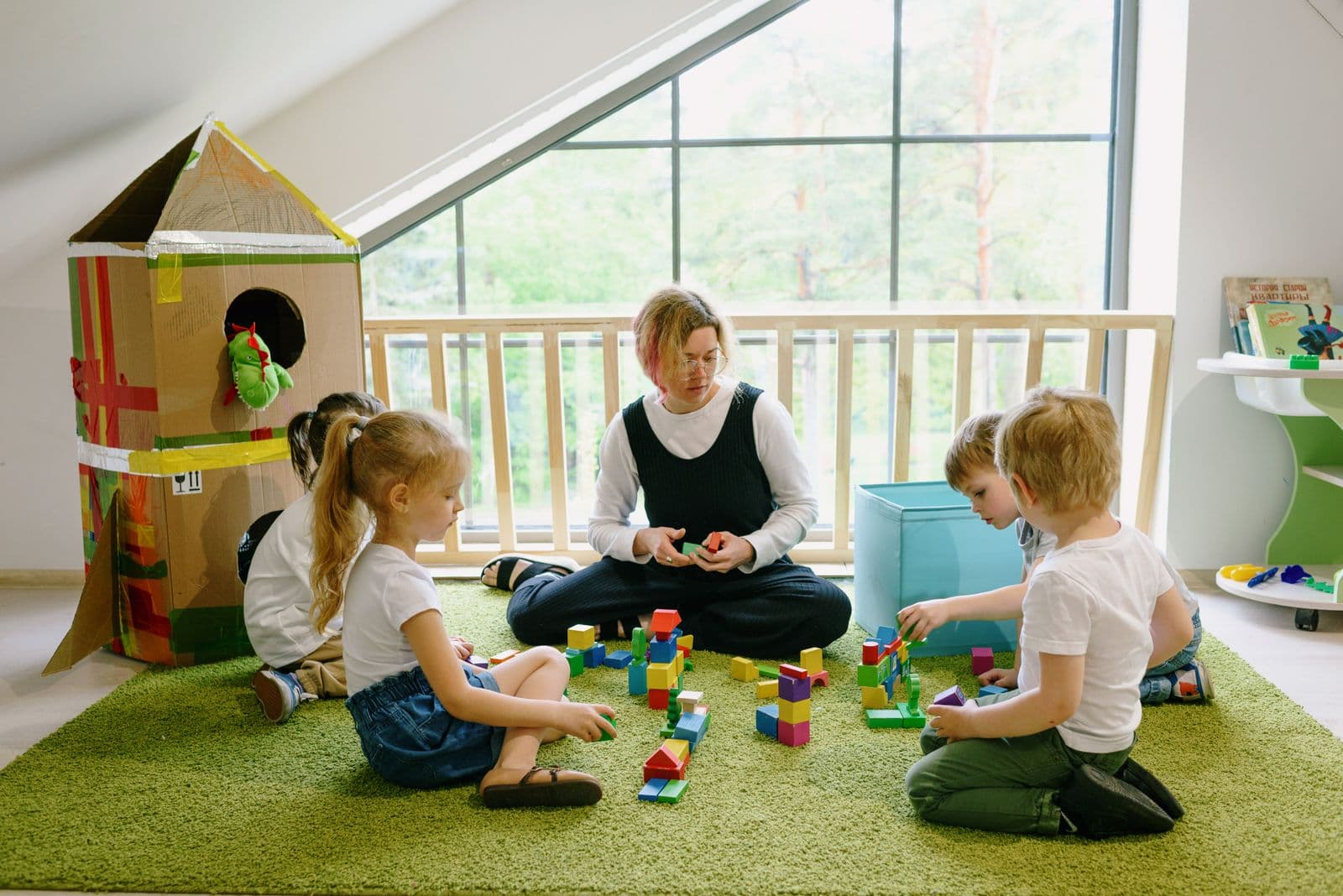 An educator sitting on the floor of a bright room with toddlers and a large cardboard playhouse.