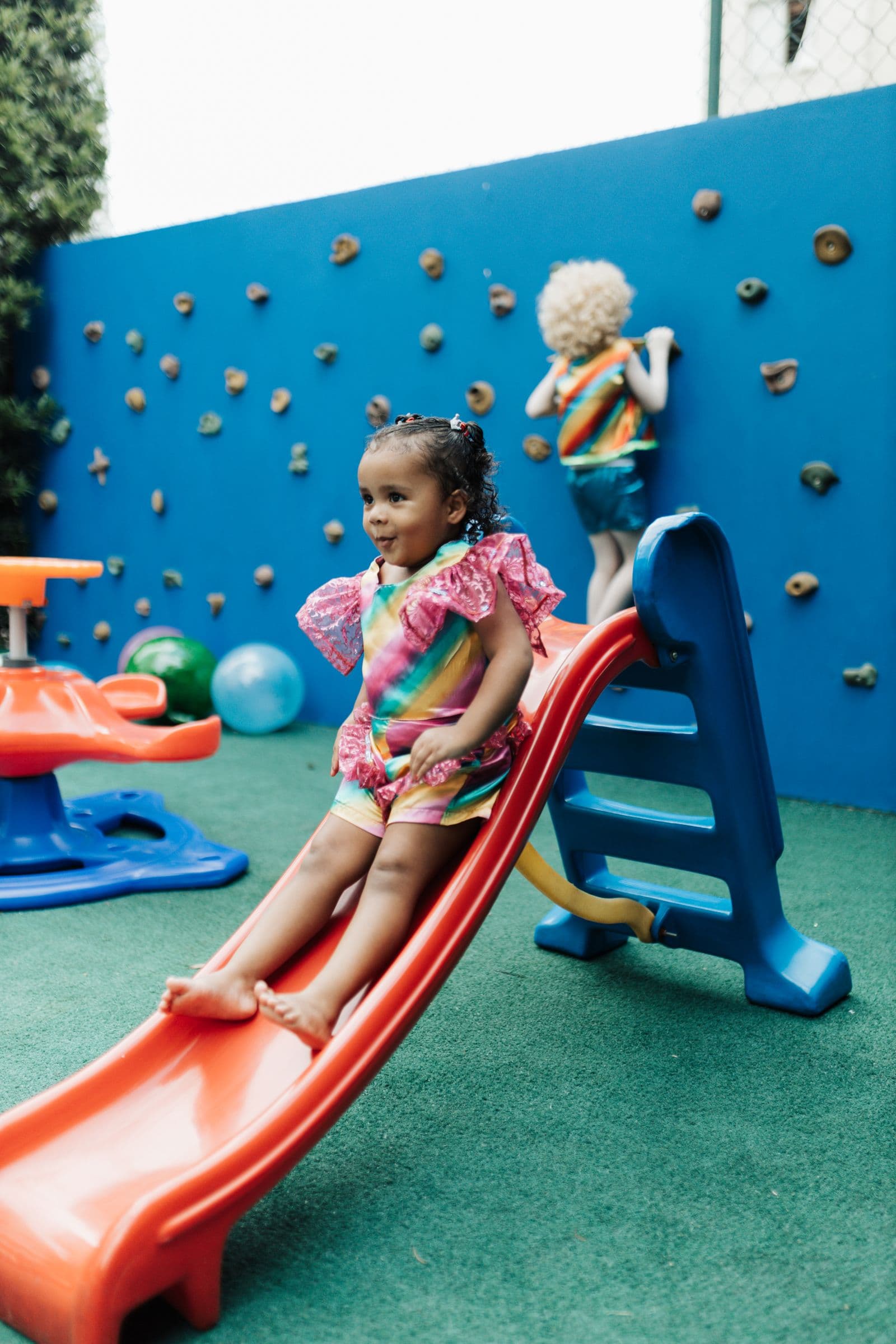 A young girl standing at the top of a small slide on an outdoor climbing frame.