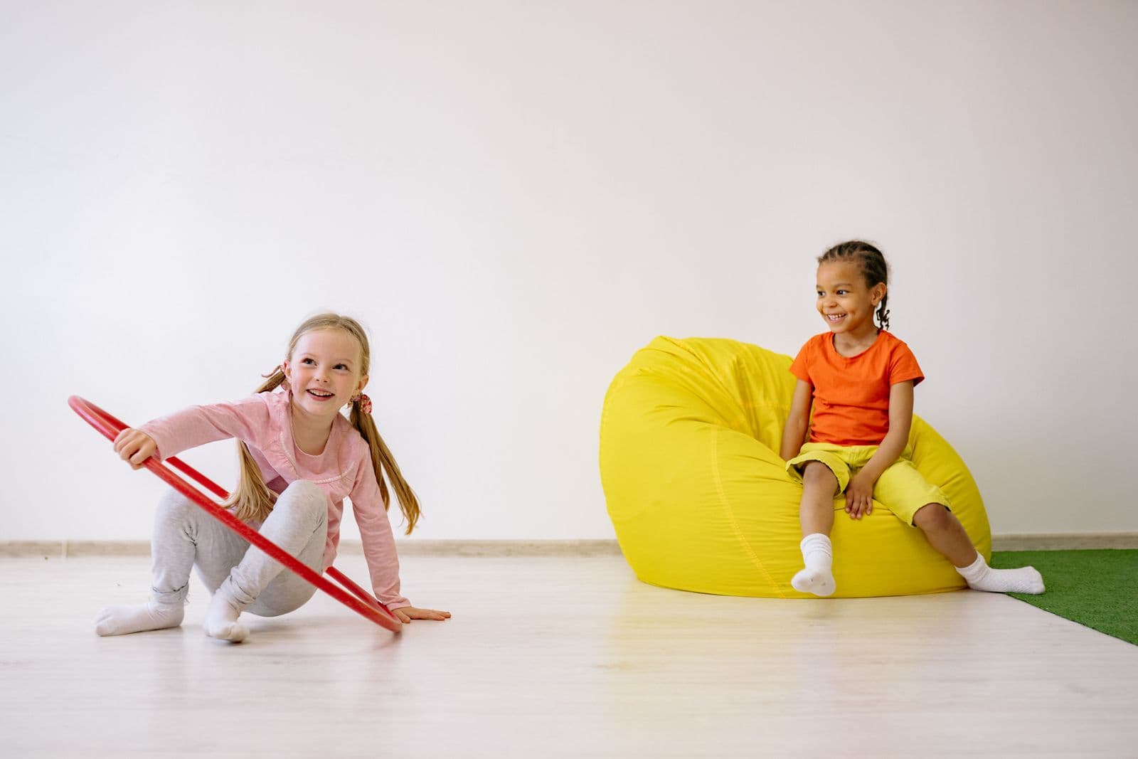 A girl playing with a hoop on the floor while another child watches from a beanbag.