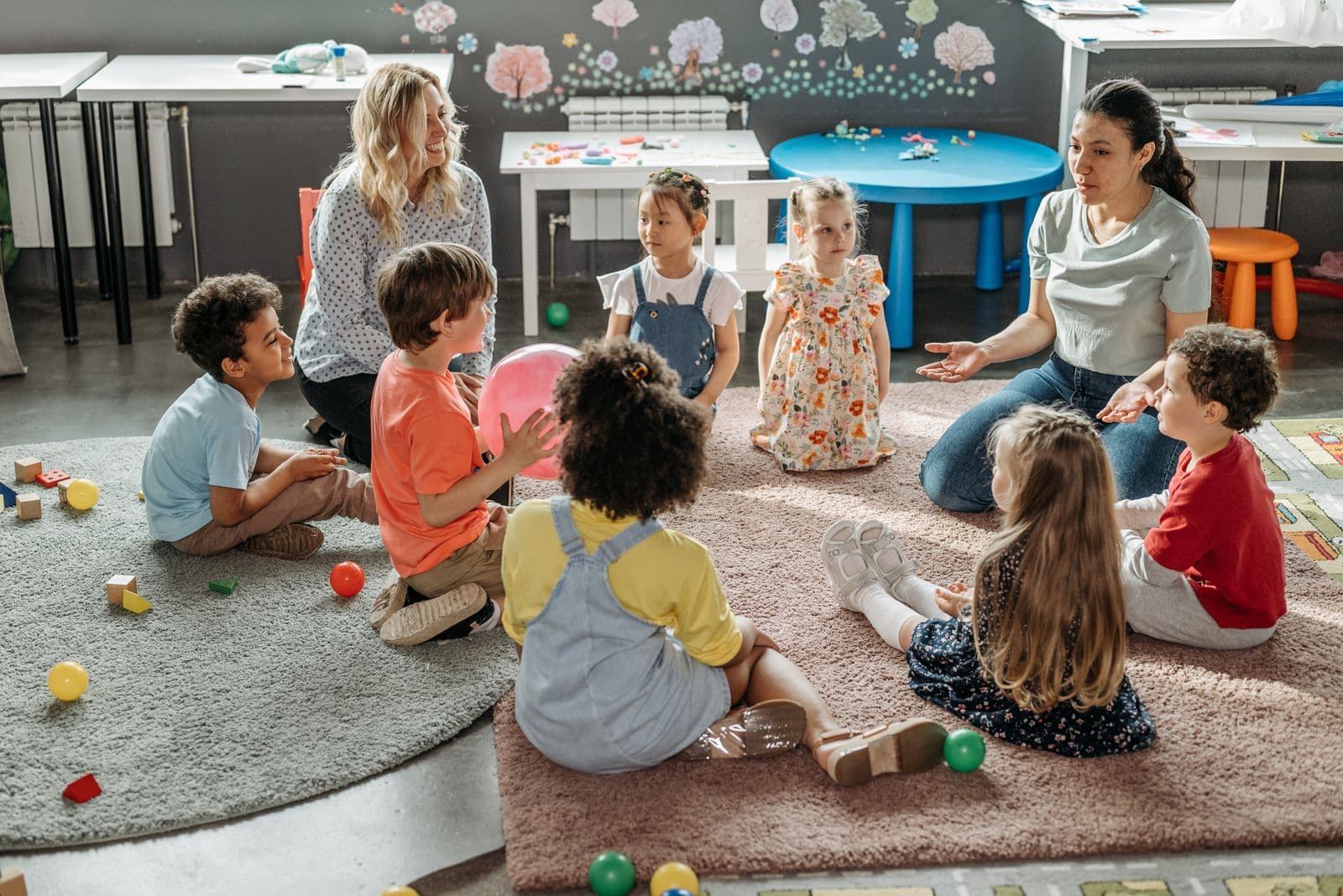 A group of children sitting in a circle on a rug with two educators, playing with soft balls.