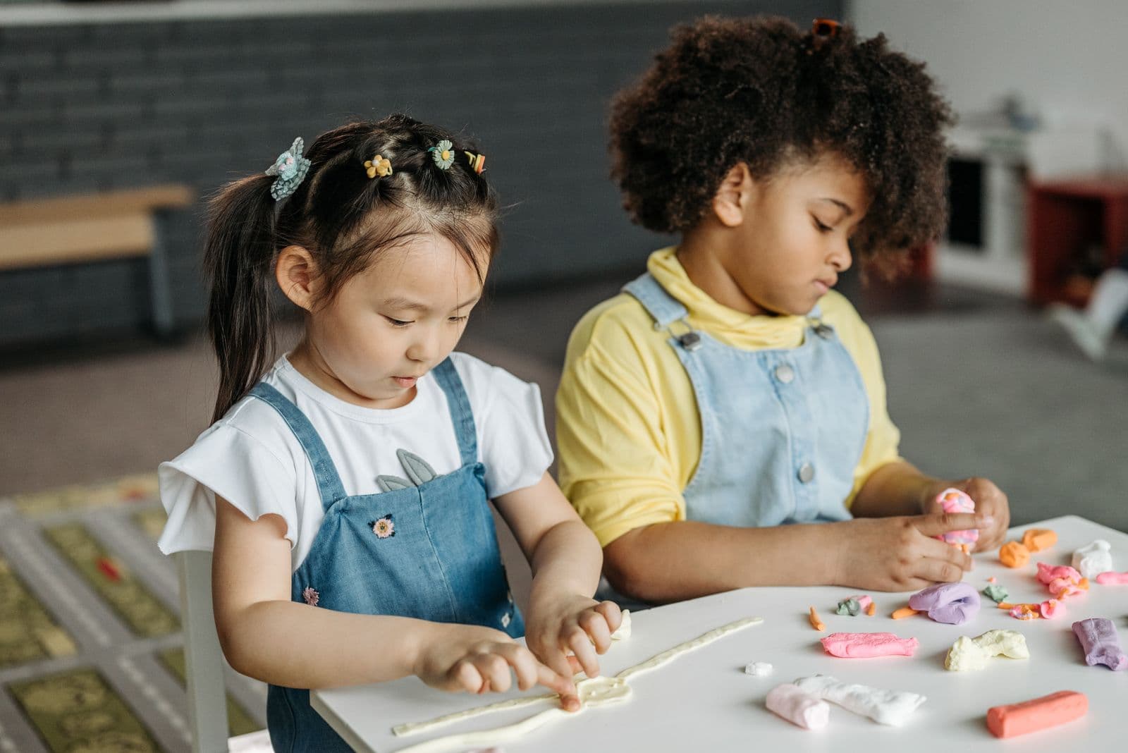 Two girls at a table working with modelling clay, one showing the other what she has made.